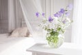 Glass vase with lilac and white floweers  in light cozy bedroom interior. White wall, bed with white linen, light blanket or plaid Royalty Free Stock Photo