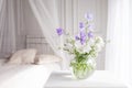 Glass vase with lilac and white floweers  in light cozy bedroom interior. White wall, bed with white linen, light blanket or plaid Royalty Free Stock Photo