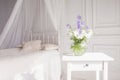 Glass vase with lilac and white floweers  in light cozy bedroom interior. White wall, bed with white linen, light blanket or plaid Royalty Free Stock Photo