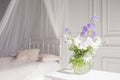 Glass vase with lilac and white floweers  in light cozy bedroom interior. White wall, bed with white linen, light blanket or plaid Royalty Free Stock Photo