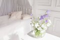 Glass vase with lilac and white floweers  in light cozy bedroom interior. White wall, bed with white linen, light blanket or plaid Royalty Free Stock Photo