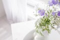 Glass vase with lilac and white floweers in light cozy bedroom interior. White wall, sunlight from window, copy space