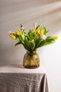 Glass vase with yellow and pink tulips on table with beige tablecloth. Gray background, diagonal shadows from wall blinds Royalty Free Stock Photo