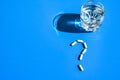 Glass of water and pills in shape of question mark on blue background. Hard light and shadows. Vitamins and prebiotics, probiotics