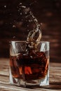 Glass glass with whiskey or cognac, frozen drink splashes from a thrown ice cube, table top made of natural aged wood, concept for Royalty Free Stock Photo