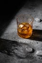 A glass of whiskey with ice on a stone gray table with ice cubes. The shadow of the glass on the table