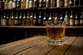 glass of whisky with ice cubes on vintage wooden table with alcohols on the shelf in background