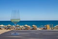 Glass of white wine on the table with shadow from palm and beach with people and umbrellas, sky and sea on the Royalty Free Stock Photo
