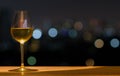 A glass of white wine on wooden table of rooftop bar with colorful bokeh of city light and space for text