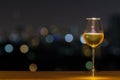 A glass of white wine on wooden table of rooftop bar with colorful bokeh of city light and space for text