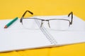 Glasses and green pencil on a white notepad. Yellow background. Close-up.