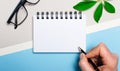 Glasses, a plant, a blank white notepad and a man`s hand with a ballpoint pen lie on a gray and light blue background. Flat lay. Royalty Free Stock Photo