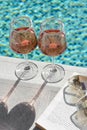 Glasses of tasty rose wine, open book and sunglasses on swimming pool edge