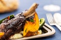 Glazed braised lamb shank on potato puree with large pumpkin and zucchini pieces in a silver tray on a blue cloth.