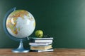 Globe, books, eyeglasses and apple on wooden table near green chalkboard, space for text. Geography lesson Royalty Free Stock Photo