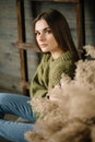 Gloomy brunette girl in a warm sweater in jeans and sneakers posing in studio with daylight. wooden background, ladder