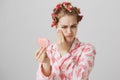 Gloomy bothered girl in pyjamas, wearing hair-curlers and holding heart-shaped mirror, wiping off makeup or mascara Royalty Free Stock Photo