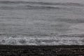 Gloomy sea in storm with smooth wave with rain drops and line of black volcanic shore, surf with foam, texture, background. Royalty Free Stock Photo