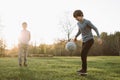 Glorious boy kicking soccer ball up, performing skills on lawn while smiling friend standing near and watching process.