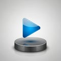 A glossy, blue, 3D play button icon is centered above a metallic, circular base. The play button Royalty Free Stock Photo