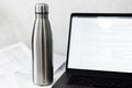 Glossy silver steel thermo bottle on desk with laptop and papers, on white background.