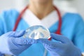 Gloved doctor holds female breast implant in his hands.