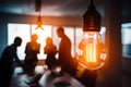 Glowing light bulb in dim room casts warm illumination on silhouettes of collaborative workspace