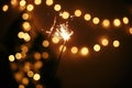 Glowing sparkler in hand on background of golden christmas tree lights, celebration in dark festive room. Happy New Year party. Royalty Free Stock Photo