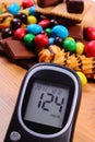 Glucose meter with heap of sweets on wooden surface, diabetes and unhealthy food