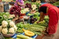 Goa, India, February 2023. Side view of barefoot Indian woman in red sari bending over stall, placing squashes zucchini.