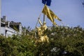 The gold Joan of Arc Maid of Orleans statue surrounded by lush green trees and plants with flags flying, buildings, blue sky