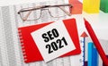 Gold-rimmed glasses, a red pen, color tables and a red notebook with a white card with the text SEO 2021 on the desktop. Business