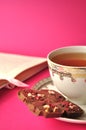 Gold and white porcelain tea cup and saucer with artisan chocolate and open book on pink background with copy space
