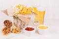 Golden colorful snacks in rustic basket and craft paper cone and sauces in bowl, lager beer on white wood table.