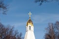 A golden cross on top of a white stone orthodox church and golden cupola surrounded by dark leafless branches of trees during a Royalty Free Stock Photo