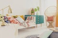 Golden lamp on nightstand in boho girl`s bedroom with colorful bedding on bed, green wooden cabinet and peacock chair with pillow