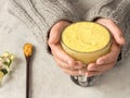 Golden milk with turmeric. Hot healthy drink in a glass bowl hold female hands. Near a wooden spoon with turmeric. Grey background