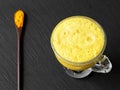Golden milk with turmeric. Hot healthy drink in a glass bowl. Near a wooden spoon with turmeric. Black stone background. Top view