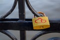 Golden Padlock With Two Red Hearts And Rusty Bracket Hangs On Black Rounded Swirls Of Bridge Fence Across River