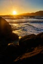 Golden sun setting behind mountains on other side of bay with rolling waves rocky shore