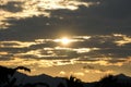 Golden sun breaking through dark, cloudy skies over a distant mountain range, with silhouetted trees in the foreground at sunrise Royalty Free Stock Photo