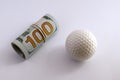 Golf ball next to a roll of hundred dollar bills of the USA with a white rubber band. The concept of sports betting, golfing