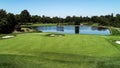 Golf course fairway, water hazard pond, sand bunkers, green with flag, lush green grass surrounded by trees against blue sky Royalty Free Stock Photo