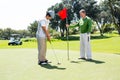 Golfer holding hole flag for friend putting ball Royalty Free Stock Photo