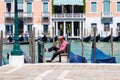 A gondolier in Venice sits looking at the phone on the waterfront.