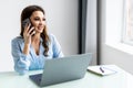 Good business talk. Cheerful young woman in glasses talking on mobile phone and using laptop with smile while sitting at her Royalty Free Stock Photo