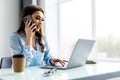 Good business talk. Cheerful young woman in glasses talking on mobile phone and using laptop with smile while sitting at her Royalty Free Stock Photo