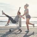 Good friends is the greatest gift. a young woman pushing her friend around on the promenade in a shopping chart.