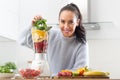 Good looking girl piles colorful mix of fruit and green leaves into a blender at home in the kitchen