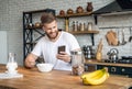 Good looking handsome bearded man is sitting in the kitchen having breakfast cereals and milk, coffee and bananas. checking his Royalty Free Stock Photo